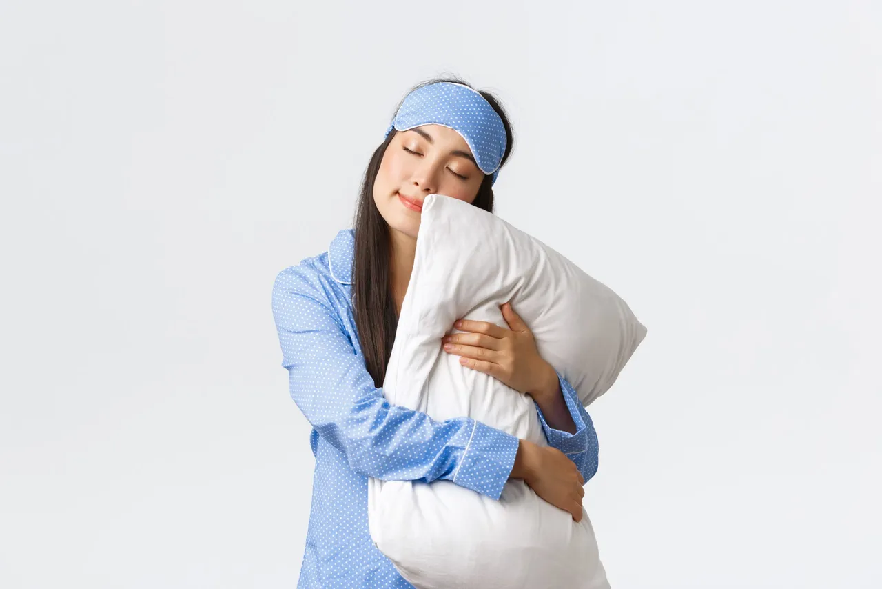 Charming sleeping asian girl in eyemask and pyjama, hugging pillow and lying on it with closed eyes, dreaming, having night sleep, posing over white background dreamy